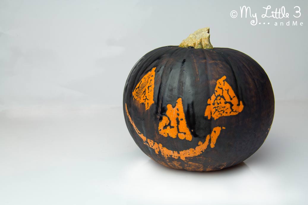 A black painted pumpkin using the wax resist method. It has a traditional carved pumpkin style face in orange where the wax has been drawn on. The rest of the pumpkin is covered in black paint.