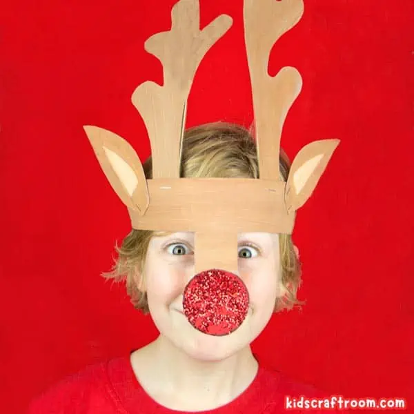 A boy in a red t-shirt wearing a reindeer hat made from paper. It has a large red nose that is covered in glitter.