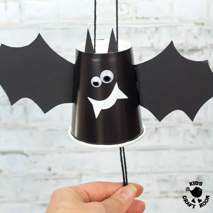 A close up of a paper cup Flying Bat Craft. A hand is pulling the strings at the bottom to make it fly.
