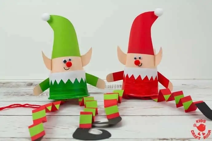 Two paper cup elf puppets sitting side by side. The one on the right is wearing red and the one on the left is wearing green.