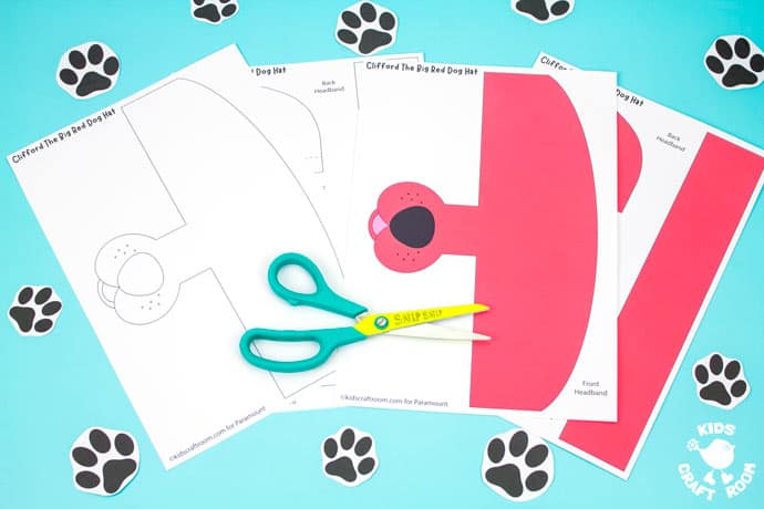 Landscape image of two Clifford The Big Red Dog hat templates lying on a blue table top surrounded by paper dog footprints.