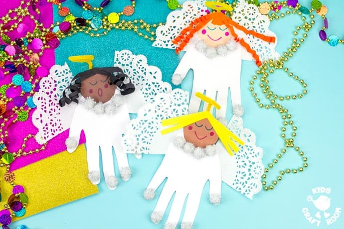 3 handprint angels with different coloured hair on a blue tabletop with colourful Christmas decorations scattered about them.