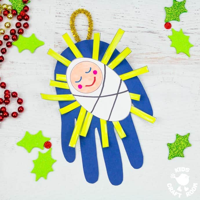 A Handprint Baby Jesus Ornament Craft lying on white table top surrounded by glittered holly leaves and red and gold Christmas bead garlands. 