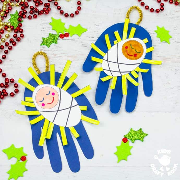 2 Handprint Baby Jesus Ornament Crafts lying on white table top surrounded by glittered holly leaves and red and gold Christmas bead garlands. 