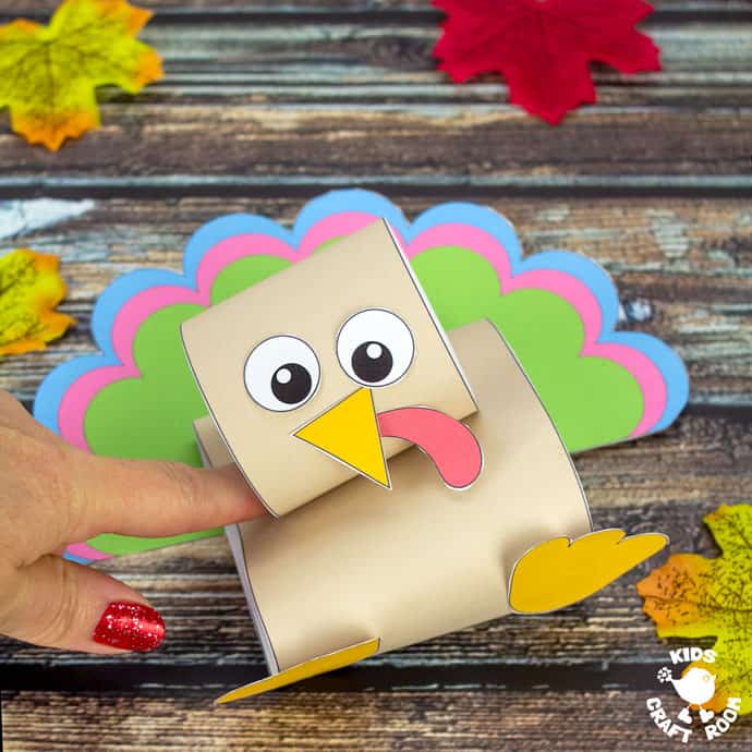 A pop up turkey craft on a wooden table top scattered with autumn leaves. A finger is pushing the turkey down.