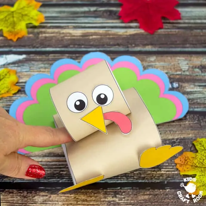 A pop up turkey craft on a wooden table top scattered with autumn leaves. A finger is pushing the turkey down.