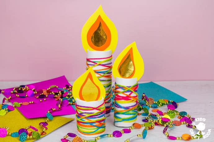 3 Cardboard Tube Candle Crafts on a table surrounded by colourful paper and Christmas decorations. 