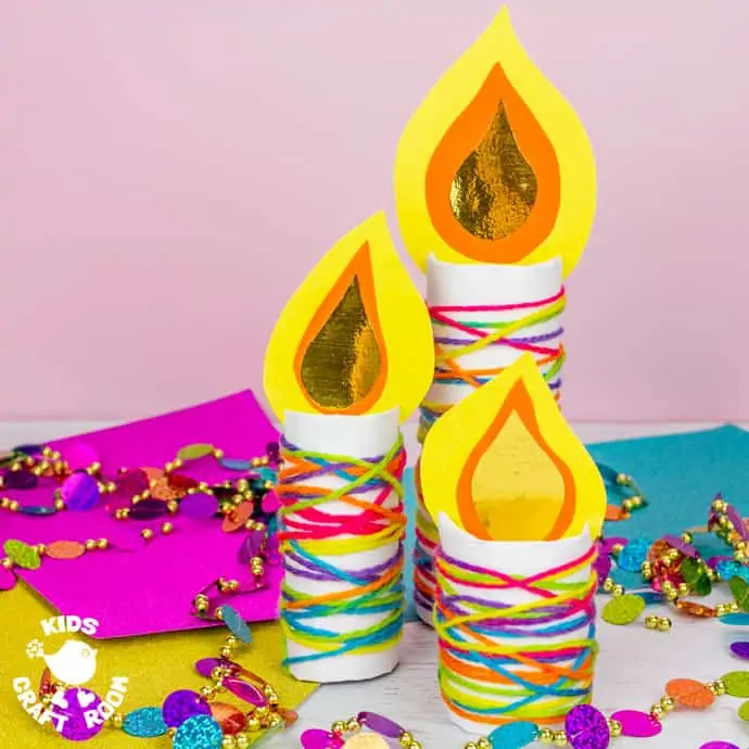A Cardboard Tube Candle Craft in 3 different sizes, on a table surrounded by colourful paper and Christmas decorations.