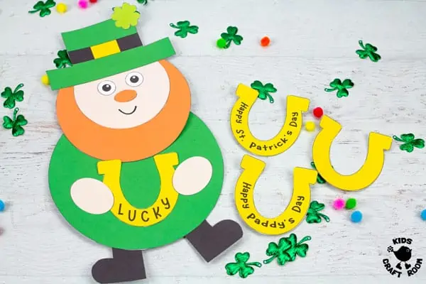 A Lucky Leprechaun craft lying on a white wood table top surrounded by scattered shamrock leaves and coloured pom poms. Next to the leprechaun are 3 lucky horseshoes.