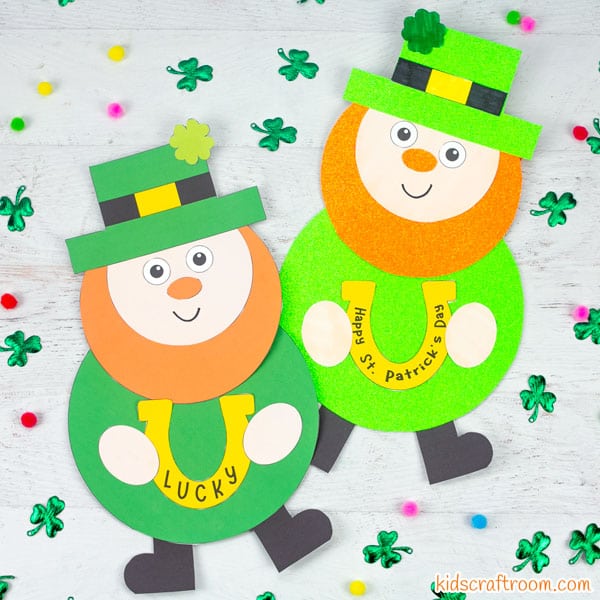 2 Lucky Leprechaun crafts lying on a white wood table top surrounded by scattered shamrock leaves and coloured pom poms.
