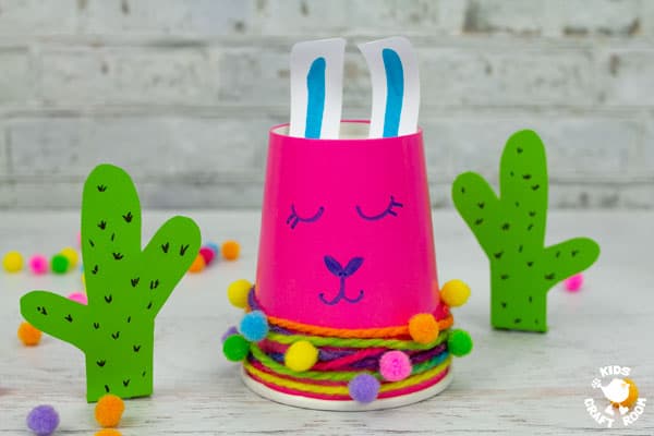 A pink paper cup llama craft with a paper cactus on either side.