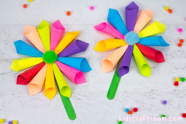 2 multicoloured sticky note flowers lying side by side on a white tabletop. Scattered about them are colourful mini pompoms.