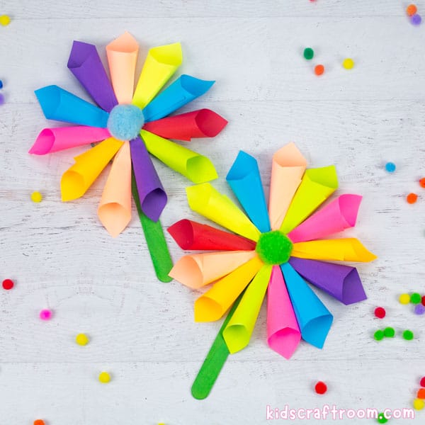 2 multicoloured sticky note flowers lying on a white tabletop. Scattered about them are colourful mini pompoms.