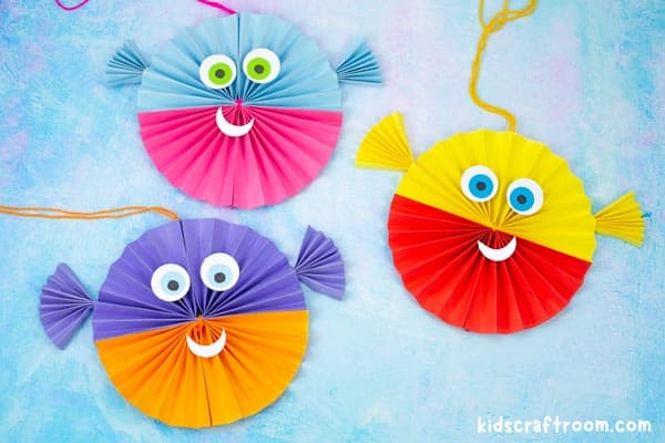 Three Paper Fan Puffer Fish Crafts lying on a blue table top. each fish has a string to hang them from and they are all different colours.