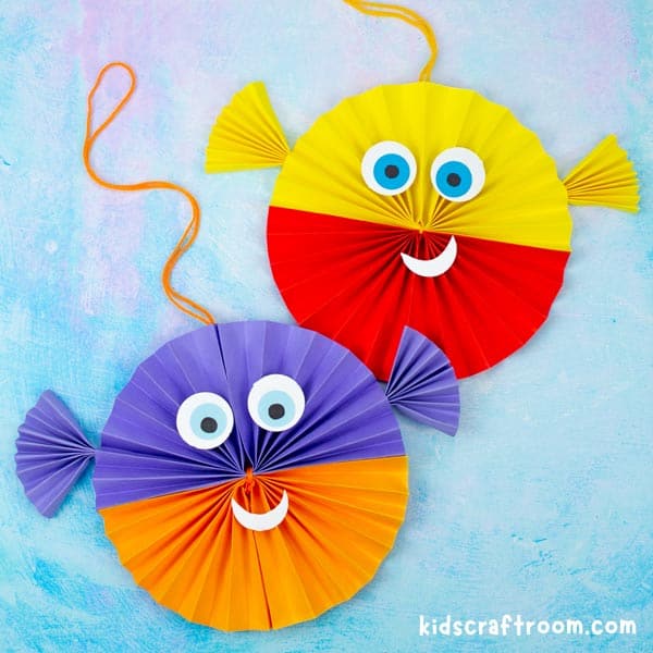 2 Paper Fan Puffer Fish Crafts on a blue background. The one on the right is purple and orange, the one on the left is yellow and red.