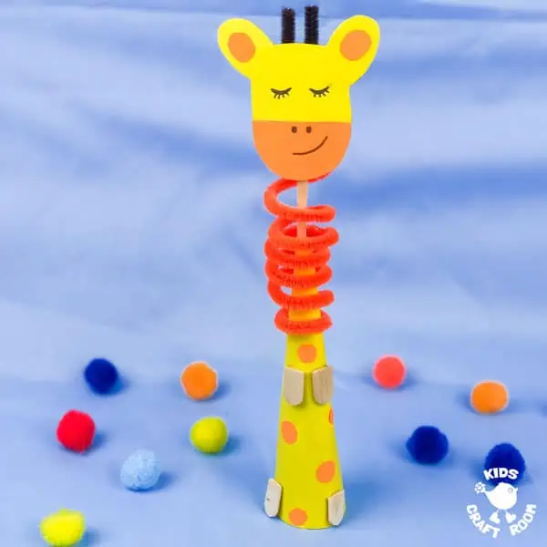 Paper Cone Giraffe Craft on a blue background with coloured pom poms scattered about.