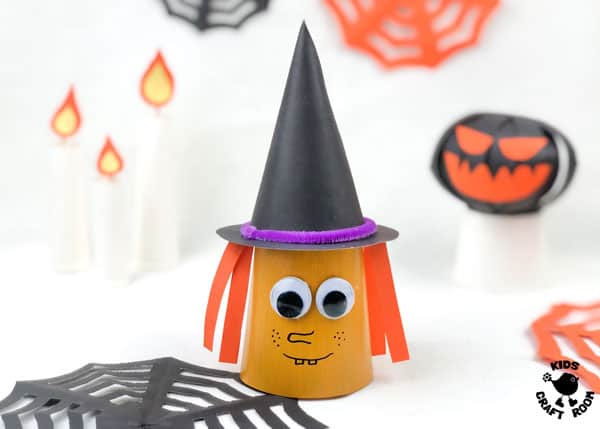 A paper cup witch sat next to a paper jack o lantern on a white table top.