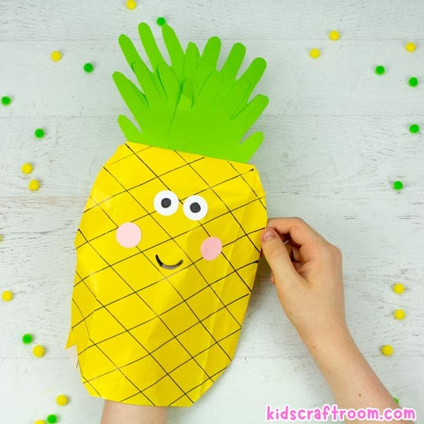 A Pineapple Paper Bag Puppet being played with over a white table top. There is a hand inside the puppet and one holding the outside.