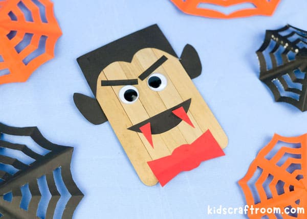 A finished Popsicle Stick Vampire Craft lying on a blue table top. It is surrounded by paper cobwebs in black and orange.