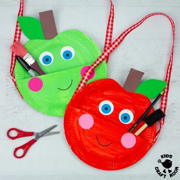 Two Paper Plate Apple Bag Crafts lying on a white surface. One is green and one is red. They have smiling faces and are holding a selection of pens, paintbrushes etc.