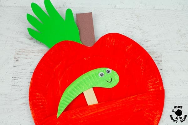 A Paper Plate Apple Puppet Craft with a green worm puppet lying on a white tabletop.