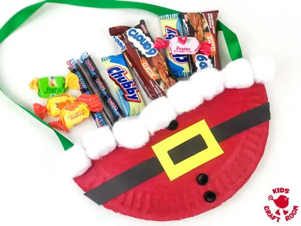 A Paper Plate Santa Treat Bag lying on a white background. The bag is full of chocolate and candy goodies.