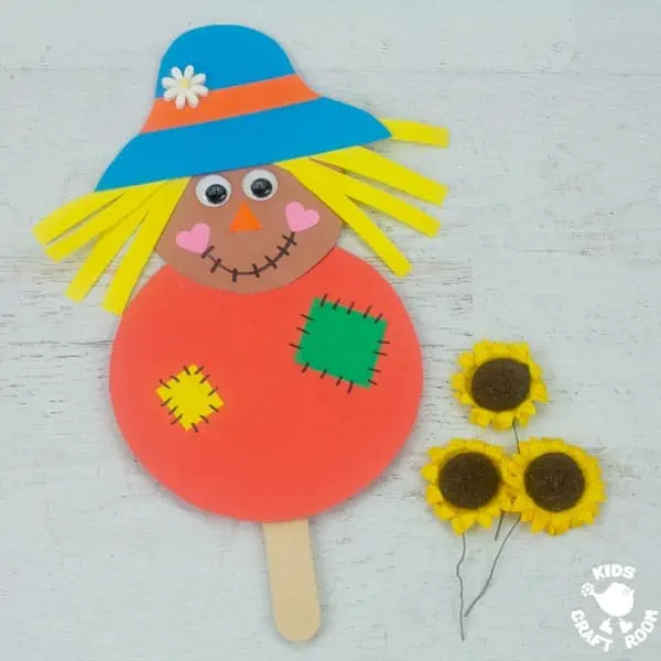A Paper Scarecrow Puppet Craft For Kids lying on a white surface. It has red trousers and a blue hat. Three mini sunflowers lie next to it.