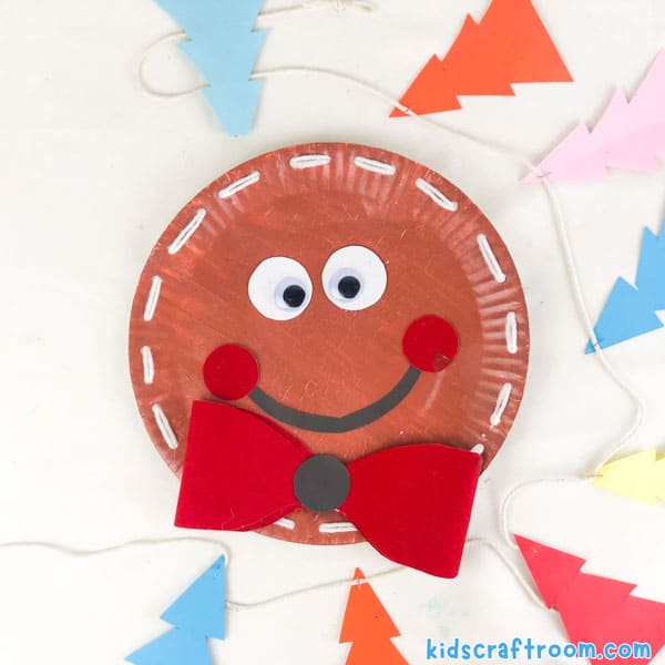 A close up of a Laced Paper Plate Gingerbread Man. He has a smiling face and a big red bow tie.