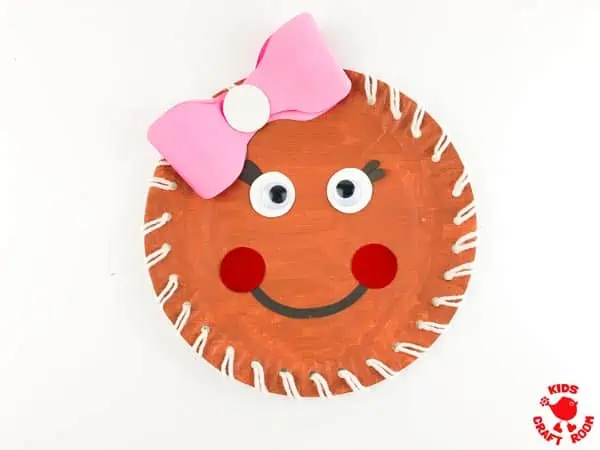 A close up of Laced a Paper Plate Gingerbread Girl. She has a big pink ribbon in her hair and a smiling face.
