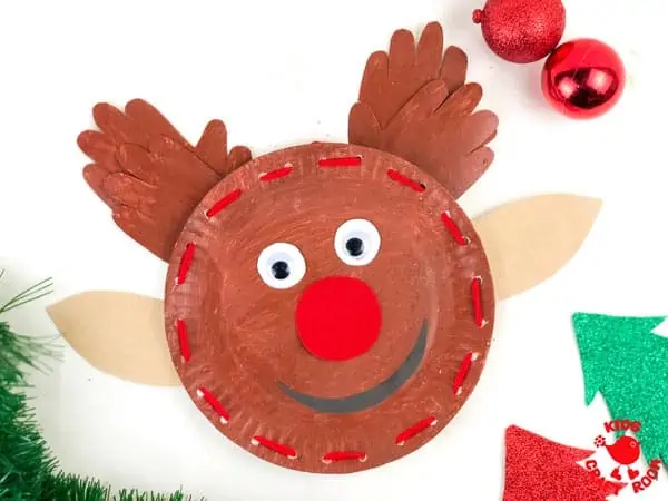 A Laced Paper Plate Reindeer with a smiling face, handprint antlers and a big red nose. It's lying on a white table top with Christmas decorations scattered around.