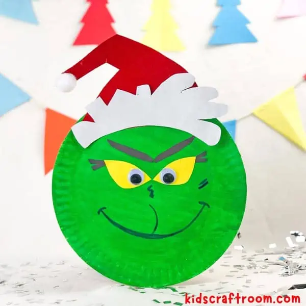 A smiling Paper Plate Grinch Craft on a white tabletop with silver glitter scattered around it.
