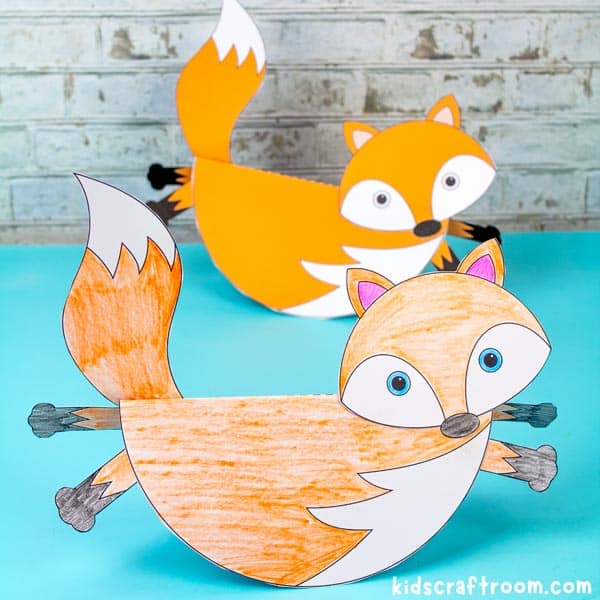 two rocking fox crafts on a blue tabletop, one behind the other. The front fox is coloured with pencils, the back is ready coloured.