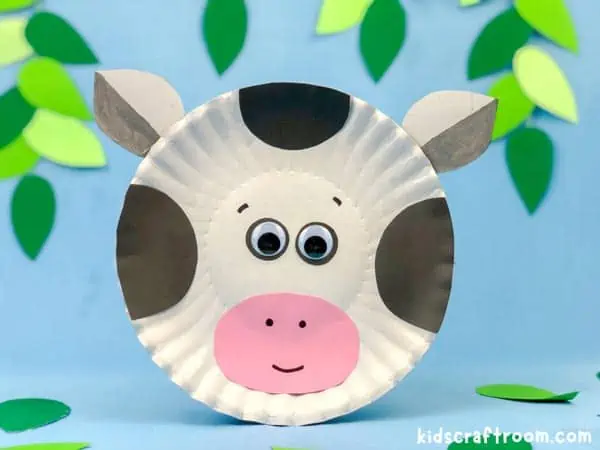 A Paper Plate Cow Craft infront of a blue background with leaves around the edges.