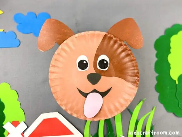 A close up of a Paper Plate Dog on a background of grass, trees, farm buildings etc cut out from coloured paper.