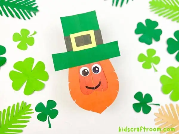 A close up of a Popsicle Stick Leprechaun Craft on a white background surrounded by paper shamrock leaves.