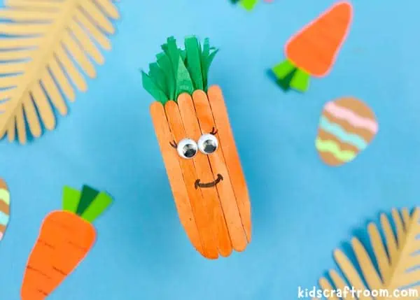 A close up of a Popsicle Stick Carrot Craft on a blue background with paper leaves and paper carrots scattered around it.