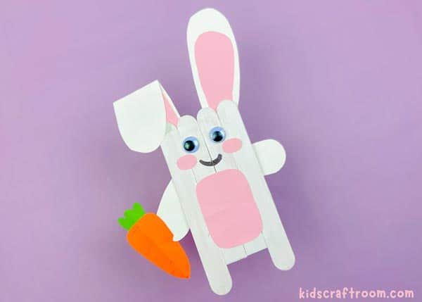 A white Popsicle Stick Easter Bunny Craft lying diagonally on a purple background. He holds a paper carrot in his right paw.