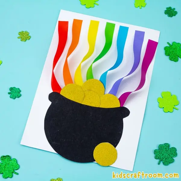 a square image showing a Rainbow Pot Of Gold Craft lying on a blue background. there are glitter shamrock leaves scattered around it.