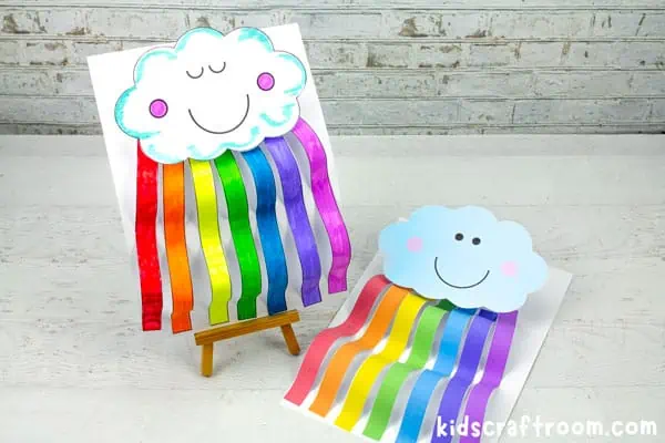 2 Rainbow Cloud Crafts. One is standing upright on a small table top easel. Its eyes are shut. another is ready coloured and lying on the table next to the first. Its eyes are open.