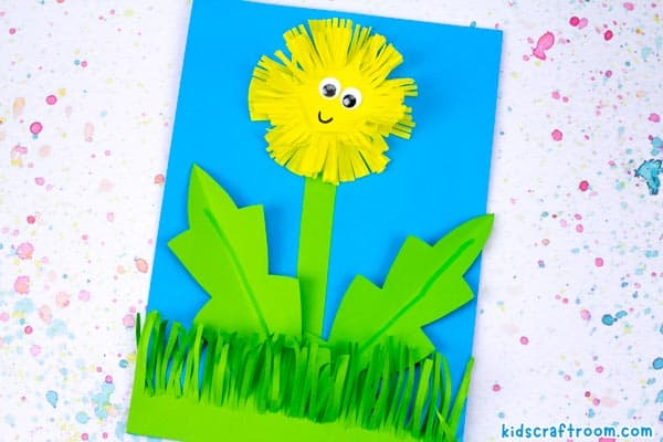 A dandelion cutting practice craft lying on a paint splattered background. It has a happy smile and wiggle eyes.