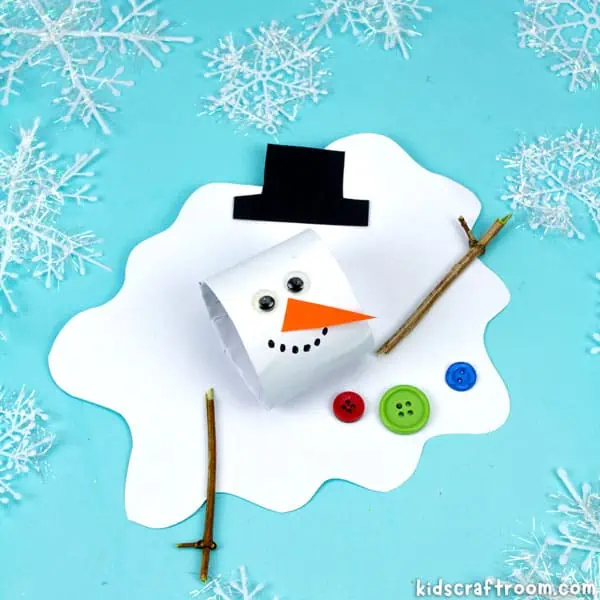 A close up of a melted snowman craft made from a white cardboard tube head and a paper body. It has wiggle eyes, a black paper hat and orange paper nose. It has twig arms and three buttons glued to its front.