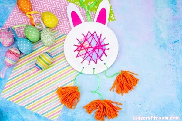 A DIY Easter Bunny Dreamcatcher lying on a blue surface with patterned papers and colored Easter eggs. The dreamcatcher is made from a white paper plate with white card ears with pink centres. The middle is woven with pink yarn and there are three orange pom-poms hanging from the bottom of it.