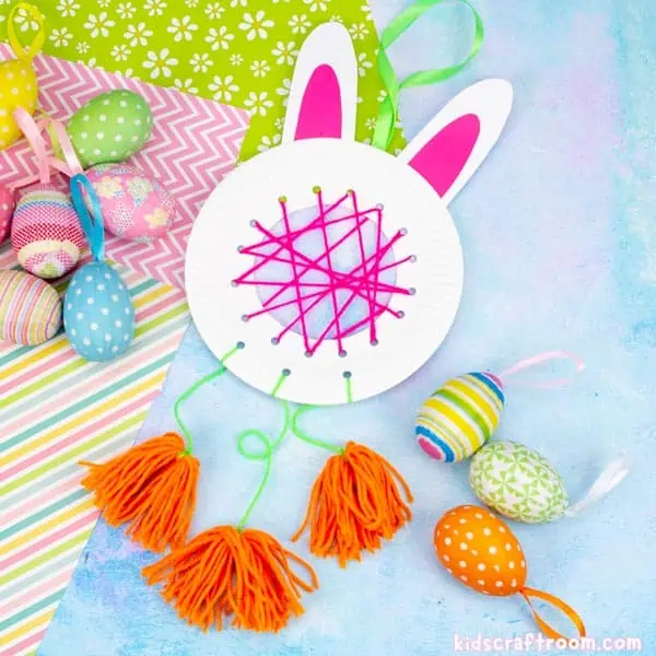 A DIY Easter Bunny Dreamcatcher lying on a blue surface with patterned papers. there are brightly colored Easter eggs scattered around it. The dreamcatcher is made from a white paper plate with white card ears. The middle is woven with pink yarn and there are orange pom poms hanging from the bottom of it.