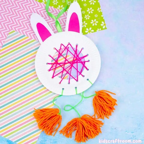 A DIY Easter Bunny Dreamcatcher lying on a blue surface with patterned pastel papers. The dreamcatcher is made from a white paper plate with white card ears. The middle is woven with pink yarn and there are orange pom poms hanging from the bottom of it representing carrots.