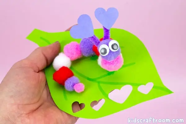 A close up of a purple Caterpillar Valentine Craft For Kids made from colored pom poms. The caterpillar has heart antennae and googly eyes. It sits on a leaf which is being held in a hand.