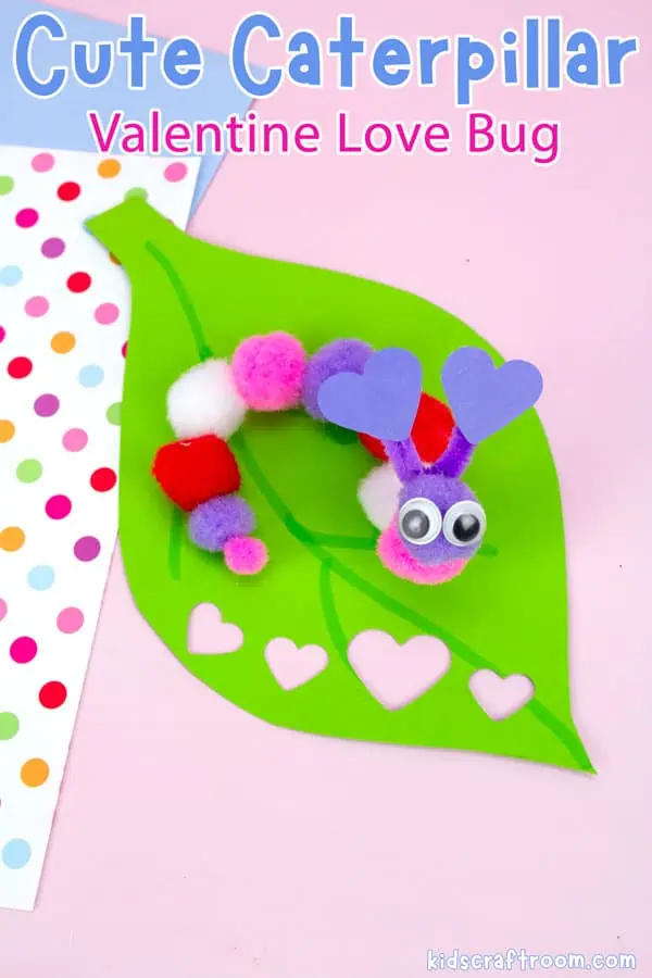 A close up of an Easy Caterpillar Valentine Craft For Kids made from colored pom poms. The caterpillar has heart antennae and googly eyes. It sits on a leaf on a pink table top.