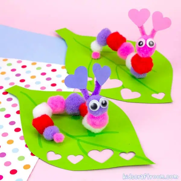 Two Easy Caterpillar Valentine Craft For Kids on green paper leaves. The caterpillar in the foreground has a purple pom pom head and the one in the background has a pink pom pom head. Their bodies are made from different coloured craft pompoms.